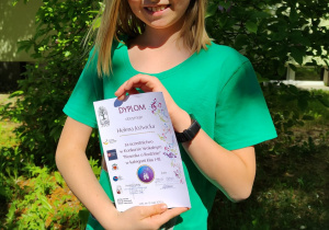 Uśmiechnięta Helenka trzymająca w ręku dyplom za udział w konkursie ,,Piosenka o Rodzinie" na tle roślinności przed budynkiem szkoły.